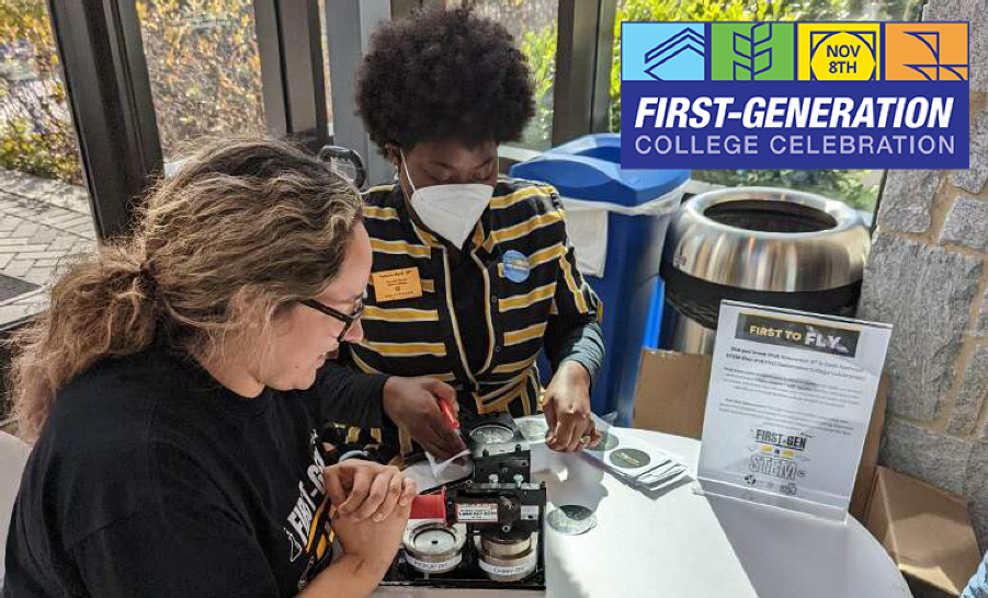 Student and staff member sitting at round table in glass enclosed space making buttons with a button maker
