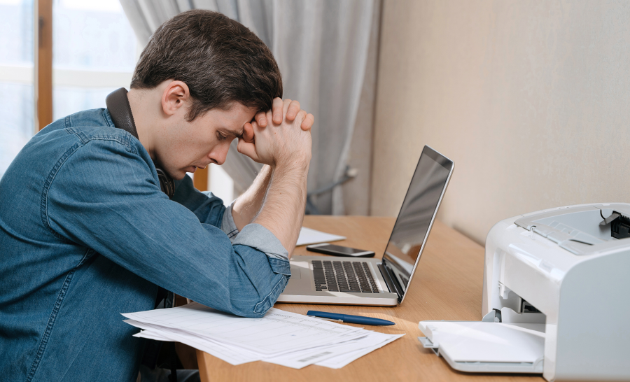 Stressed male student holds head while seated at desk with laptop computer