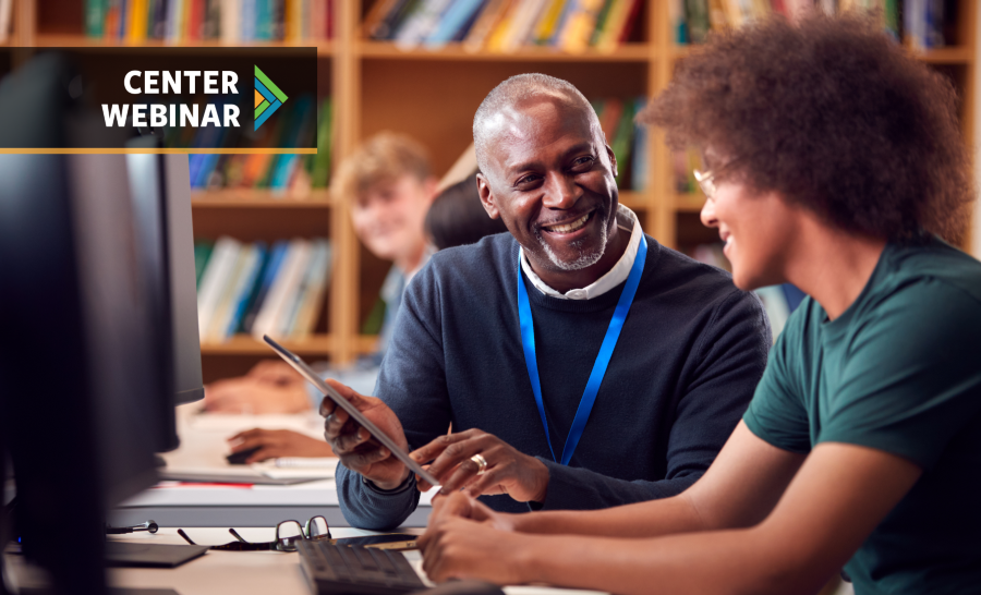 Black male student working at desktop computer in library alongside black male professor wearing a blue lanyard and gesturing at a tablet